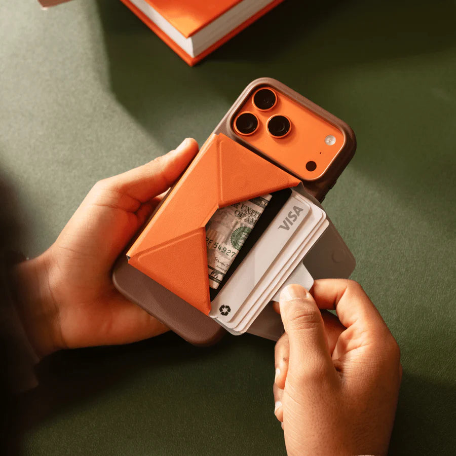A person holds an orange and gray smartphone wallet case with an orange cover, showing card slots with cards partially inserted, including a Visa card and cash. The background is a dark green surface with a blurred orange book in the top corner. Holding a moft travel wallet